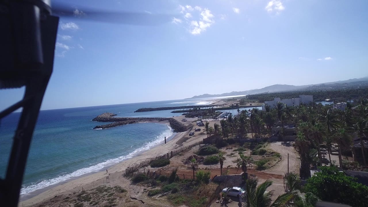 Wide elevated frame of Playita showing the curved beach, turquoise and deep blue water, palm grove, beach road, low buildings, distant coastline, and a dark rig shape near the top left.