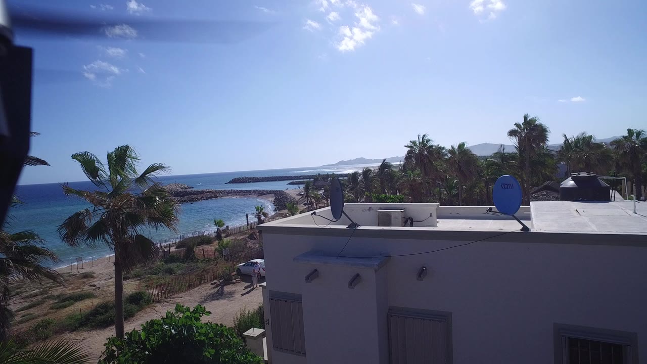 Elevated view over a white coastal building, palm trees, ocean, and blue sky.