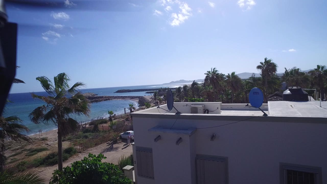 At 0:01, the Playita coastal frame keeps the white building and rooftop dishes in front while palm trees, a dirt road, blue ocean, rock outcrops, and no visible people fill the background.