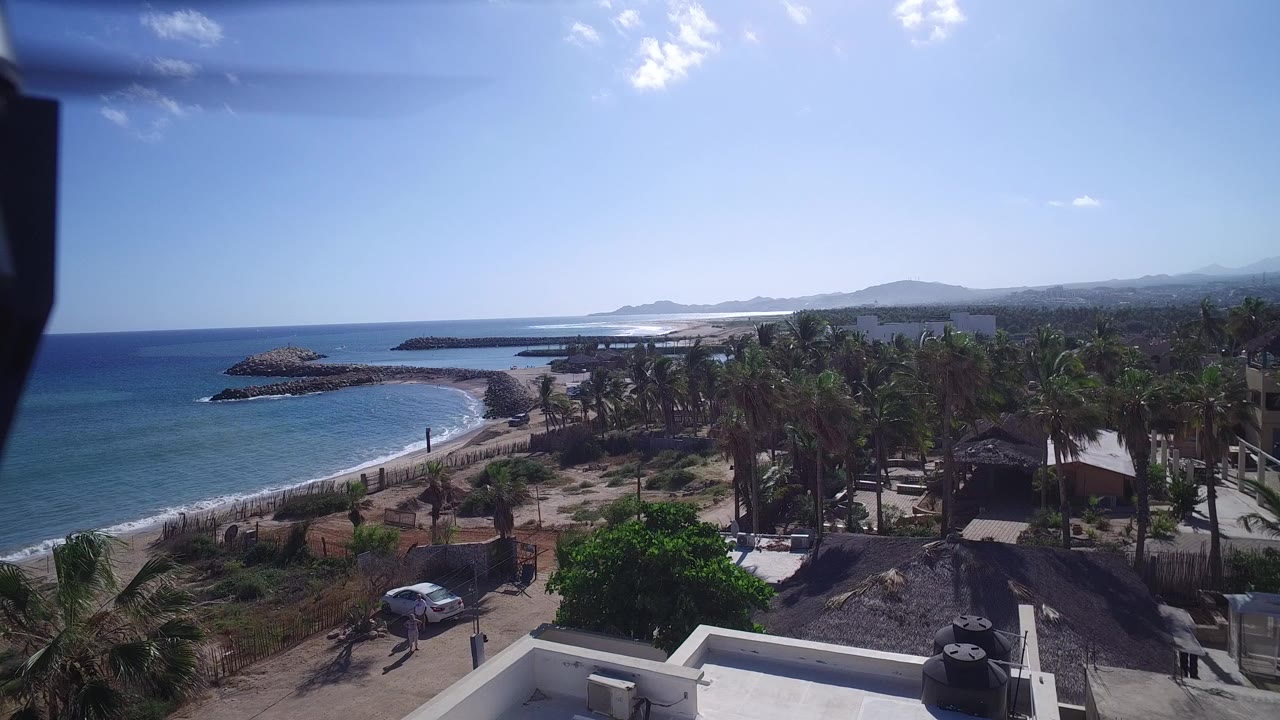 Aerial view over Playita, Baja California Sur, Mexico, with a curved beach, rock breakwaters, palms, low buildings, distant hills, and no visible people.