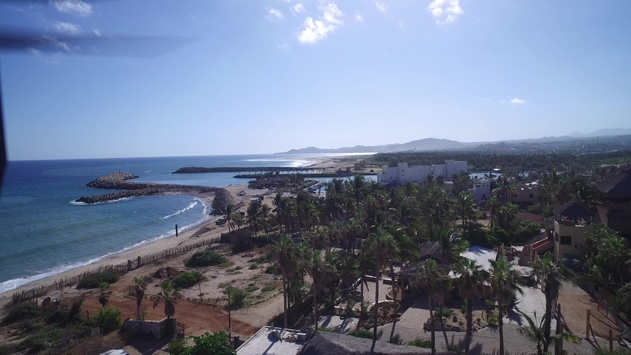 Aerial frame over Playita, Baja California Sur, showing blue ocean, a sandy beach, palm-covered development, roads, white buildings, distant hills, and no visible people.