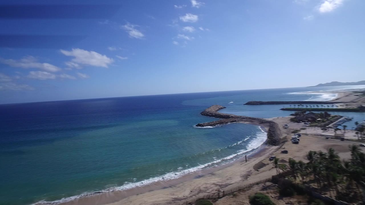 High aerial view over Playita, Baja California Sur, with turquoise ocean, a curving sandy beach, rock breakwaters, distant shoreline, and tiny beachgoers visible near the surf.