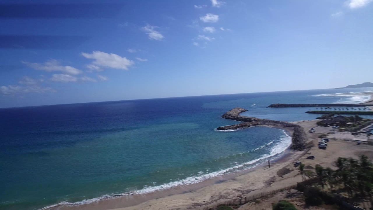 Aerial frame over Playita shows the shoreline from a slightly shifted angle, with dark blue offshore water, turquoise shallows, sandy beach, rock breakwaters, and small figures near the waterline.