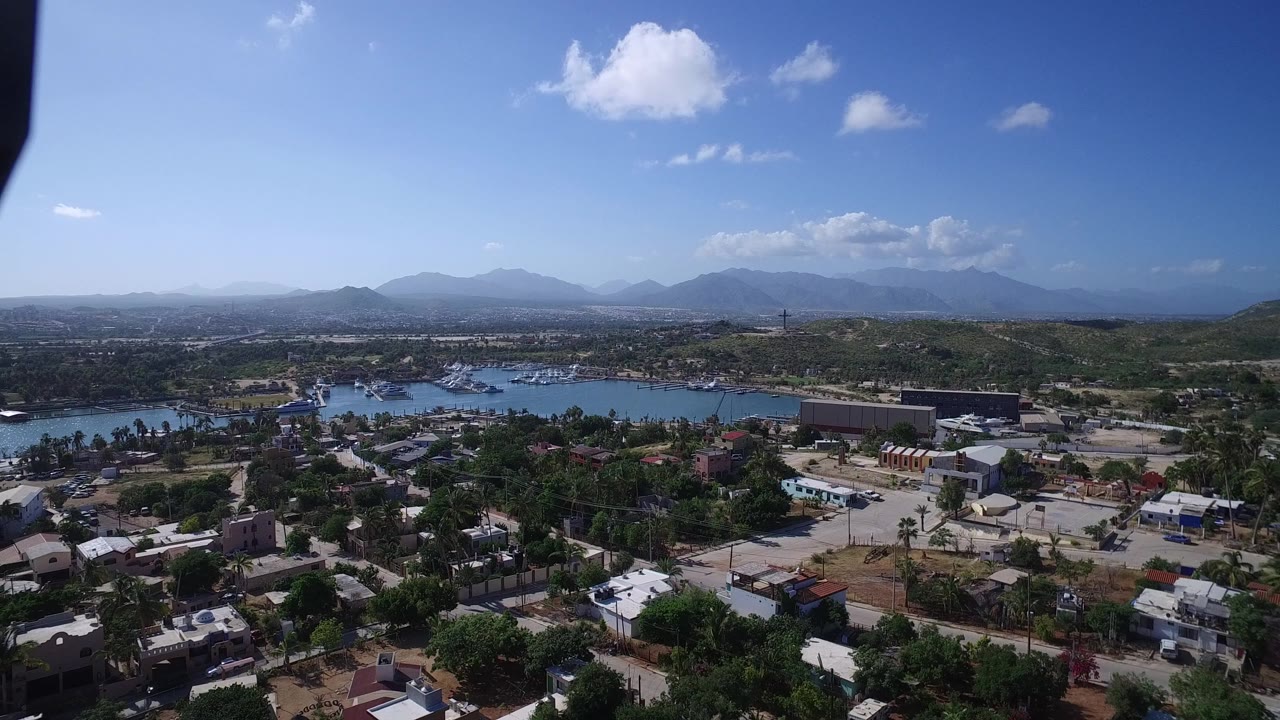 Wide drone-height view of Playita, Baja California Sur showing the bay, clustered buildings, roads, low green hills, hazy mountains, and no visible people.