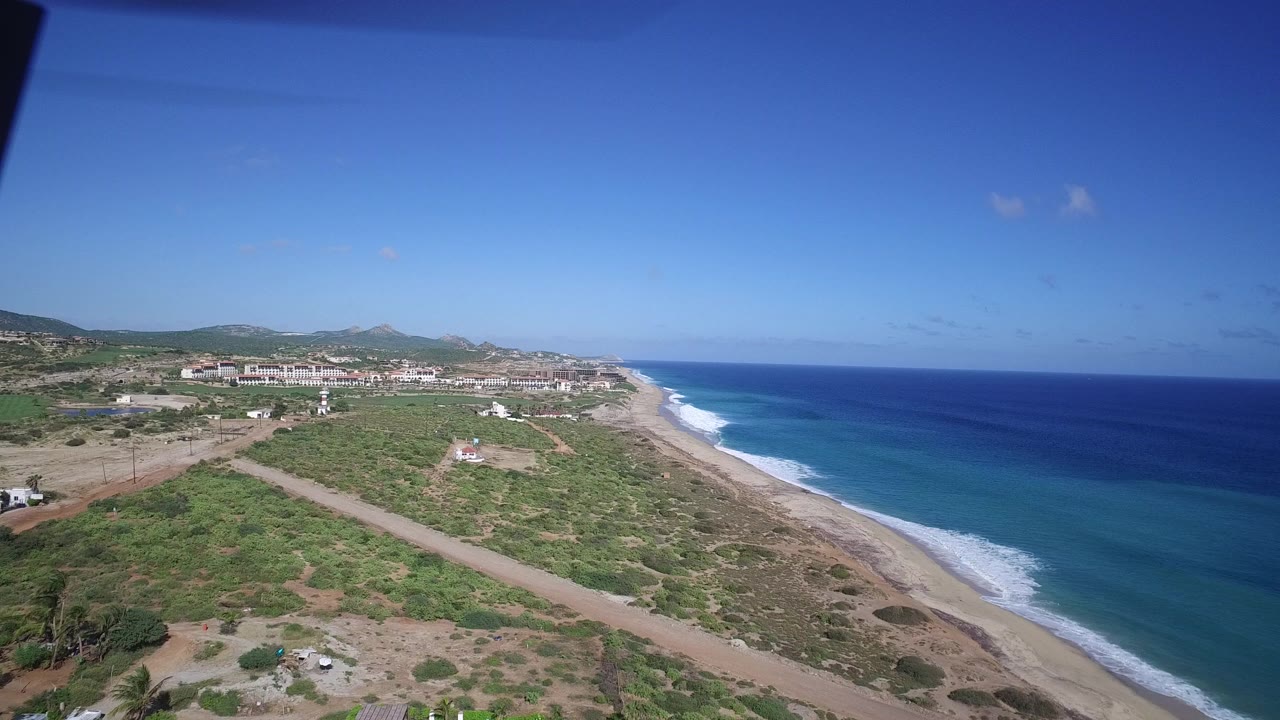 Aerial coastline with beach, turquoise ocean, scrubland, road, and distant hills.