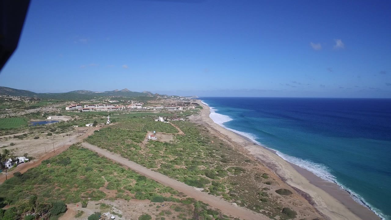 Aerial coastline with sandy beach, blue ocean, scrubland, and distant buildings.