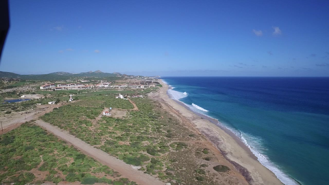 Aerial view over Playita, Baja California Sur, with blue ocean on the right, surf on a pale beach, scrubland and dirt roads inland, distant buildings and low hills, and no visible people.