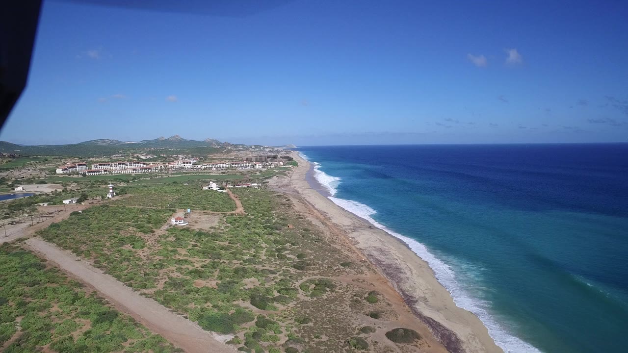 High aerial frame of Playita coastline showing scrub vegetation, dirt tracks, distant settlement, a diagonal beach, white surf, broad blue water, and no visible people.
