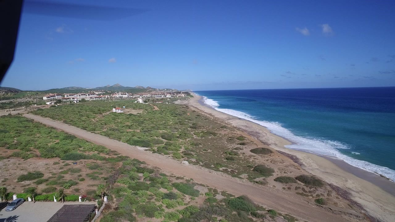 Elevated coastal view with beach, blue ocean, scrubland, road, and distant buildings.