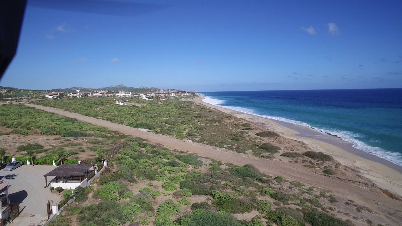 Early wide frame of the Playita coastline showing surf bands along the beach, green scrubland around a dirt road, white buildings near the horizon, and no visible people.
