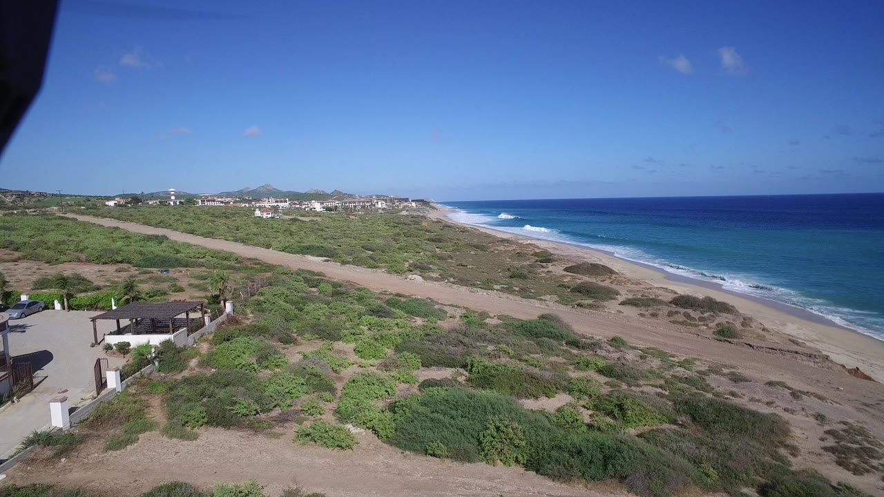 Aerial view of sandy beach, green scrubland, blue ocean, and distant buildings.