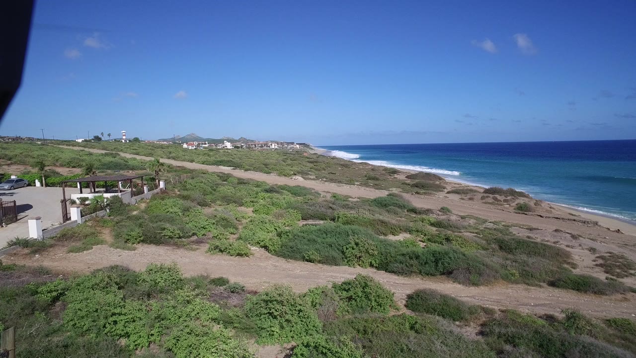 Opening view over Playita, Baja California Sur, Mexico with green coastal scrub, sandy paths, white structures at left, blue ocean at right, and no visible people.