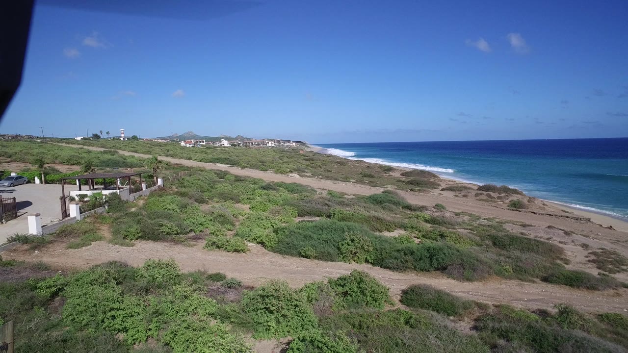 A high coastal view in Playita shows sunlit dunes and shrubs leading toward a blue sea with small surf lines, distant buildings, and no visible people.