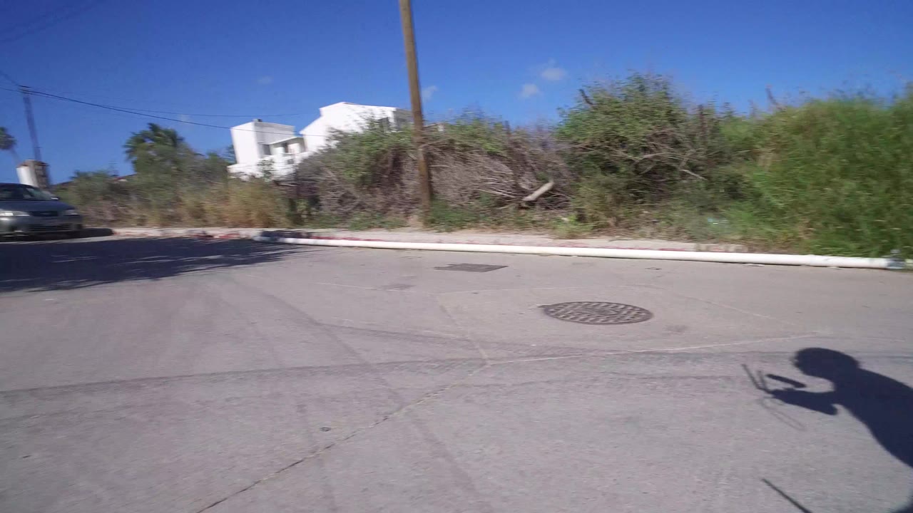 Road and curb in Playita, Baja California Sur with tall green brush, a white building in the distance, a parked or passing car at left, and no visible people.
