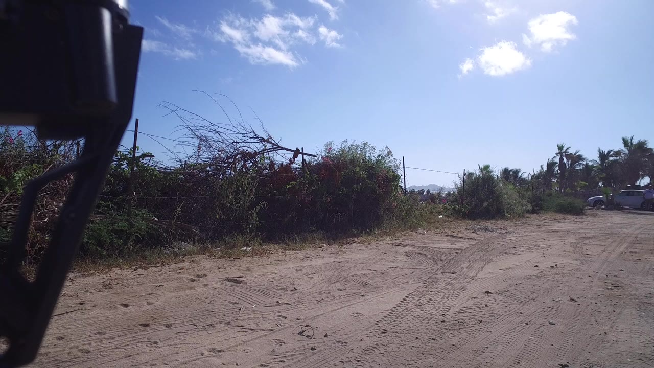 A sandy road in Playita, Baja California Sur runs past brush and wire fencing under a bright blue sky, with a dark camera part at the left edge and no visible people.