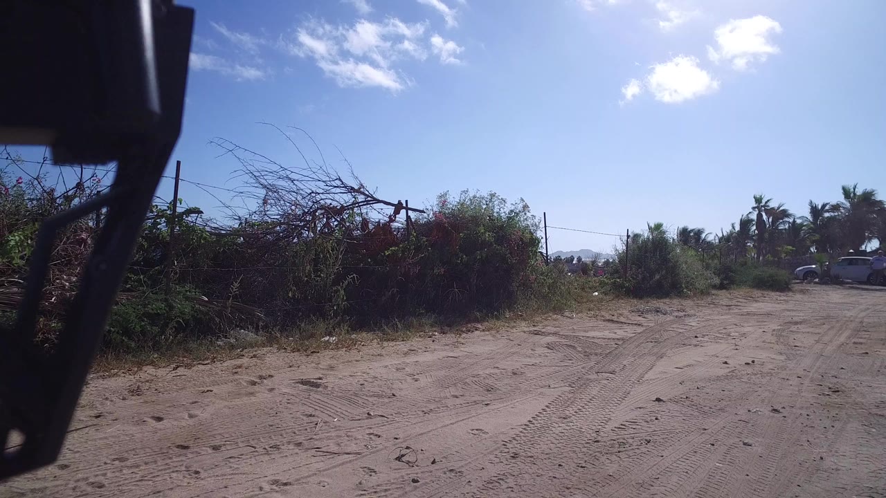 The Playita road remains framed by dry brush and fencing, with palms and a light-colored parked vehicle in the distance and no visible people.