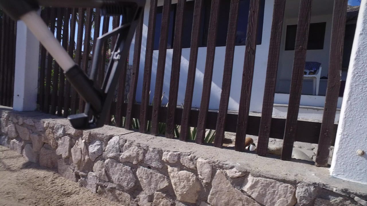 Low view of dark wooden gate bars and a stone base wall at a white house entrance in Playita, Baja California Sur, with no visible people.