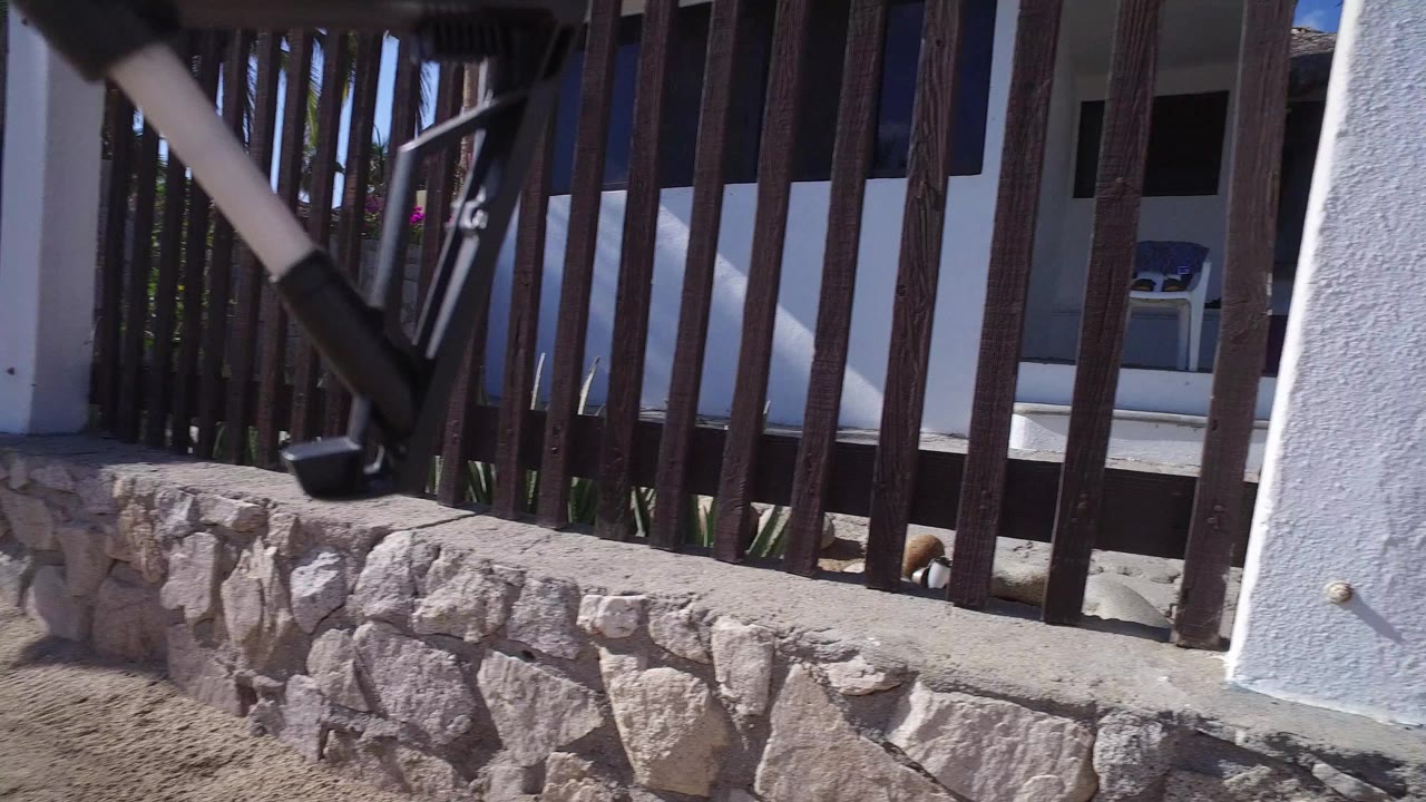 Closer view through vertical wooden gate bars toward the white Playita house entry, stone wall, shaded porch, and a small light-colored object near the base, with no visible people.