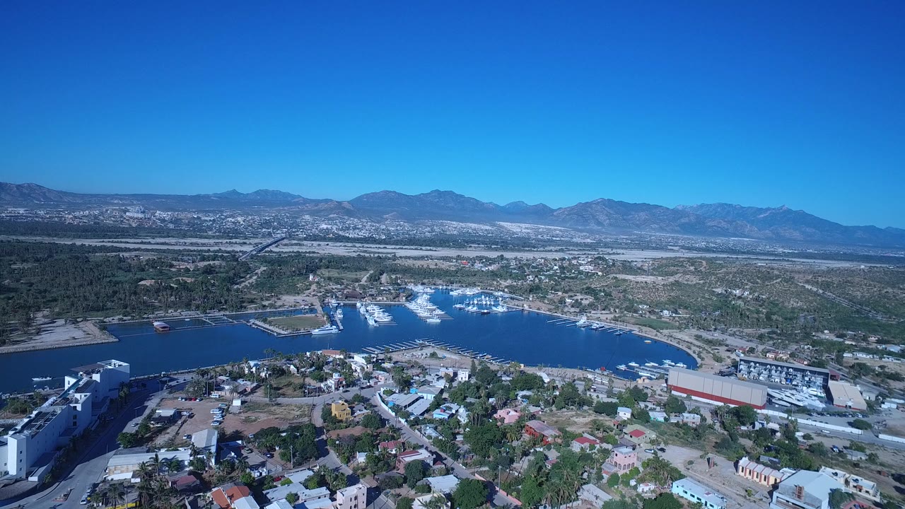Aerial frame over Playita with the harbor basin centered beyond residential streets, palms and low buildings in the foreground, and mountains across the horizon with no visible people.
