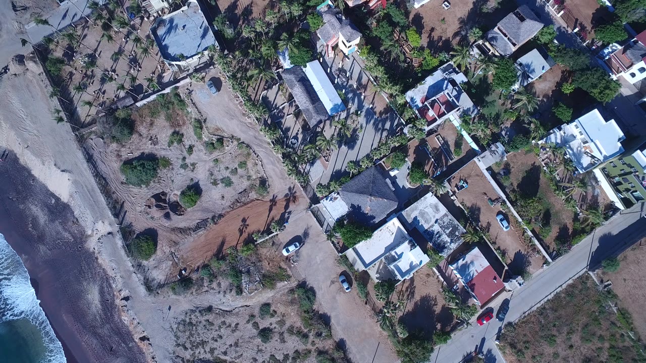Overhead frame of Playita homes and palms arranged along narrow roads, with sandy open ground and a sliver of breaking surf at the left, no visible people.
