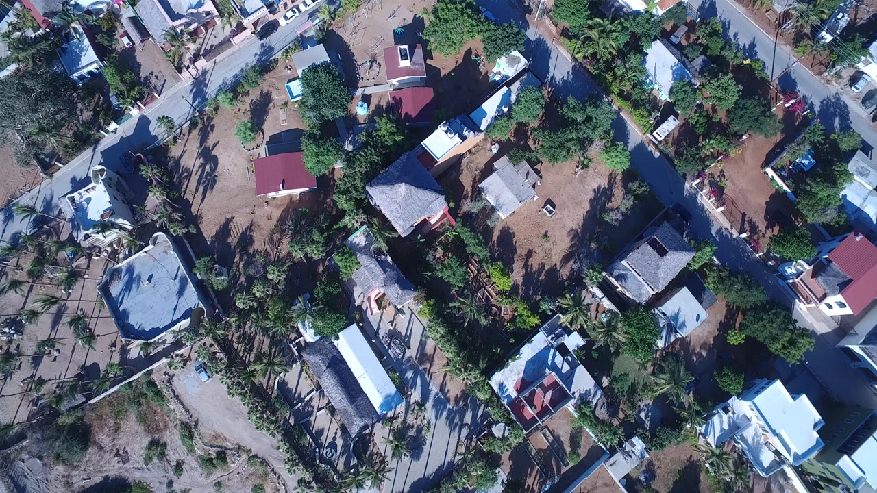Drone view over Playita, Baja California Sur with clustered homes, a red-roofed building, pale roads, dense greenery, and no visible people.