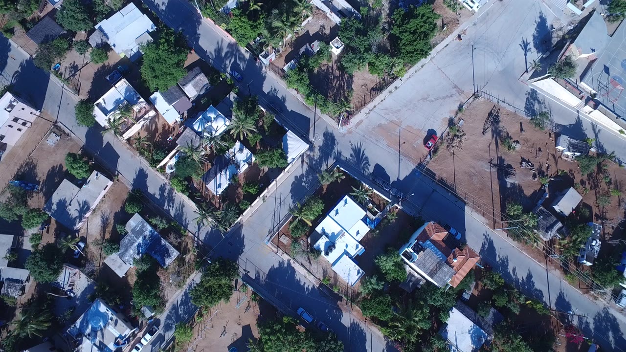 Aerial frame over Playita, Baja California Sur, with a diagonal concrete road, clustered homes, shaded trees, dry yards, and no visible people.