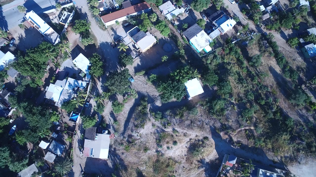 Overhead frame from Playita, Baja California Sur, showing scattered low buildings, trees, dirt paths, and a paved road near the waterfront, with no visible people.