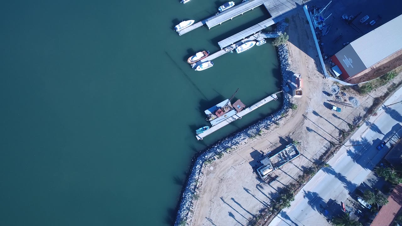 High overhead view in Playita, Baja California Sur, Mexico, showing green-blue water on the left, docked small boats, rock shoreline, dirt access area, and no visible people.