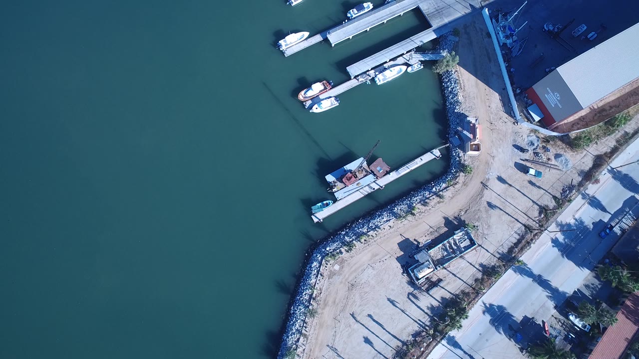 Overhead Playita marina frame with broad green-blue water, several small boats tied to docks, a curved rock edge, dirt shoreline access, and no visible people.