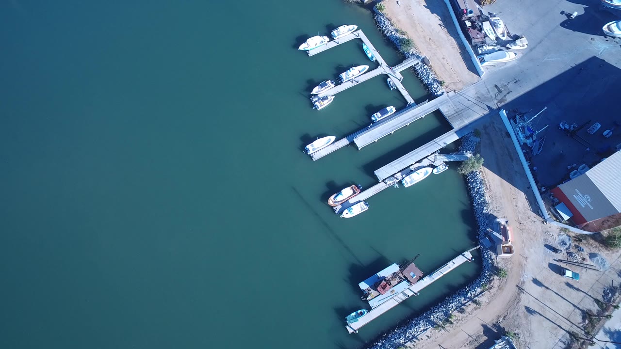 Overhead frame of Playita, Baja California Sur harbor water with two dock sections, small white boats, rocky shoreline, paved area, and no visible people.
