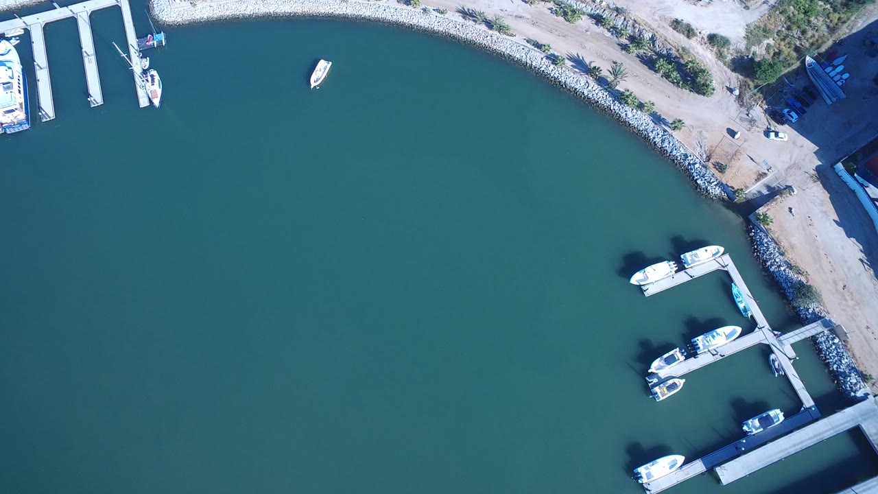 High aerial frame over Playita, Baja California Sur, Mexico, showing teal water, a curved rock shoreline, marina docks, several boats, and no visible people.