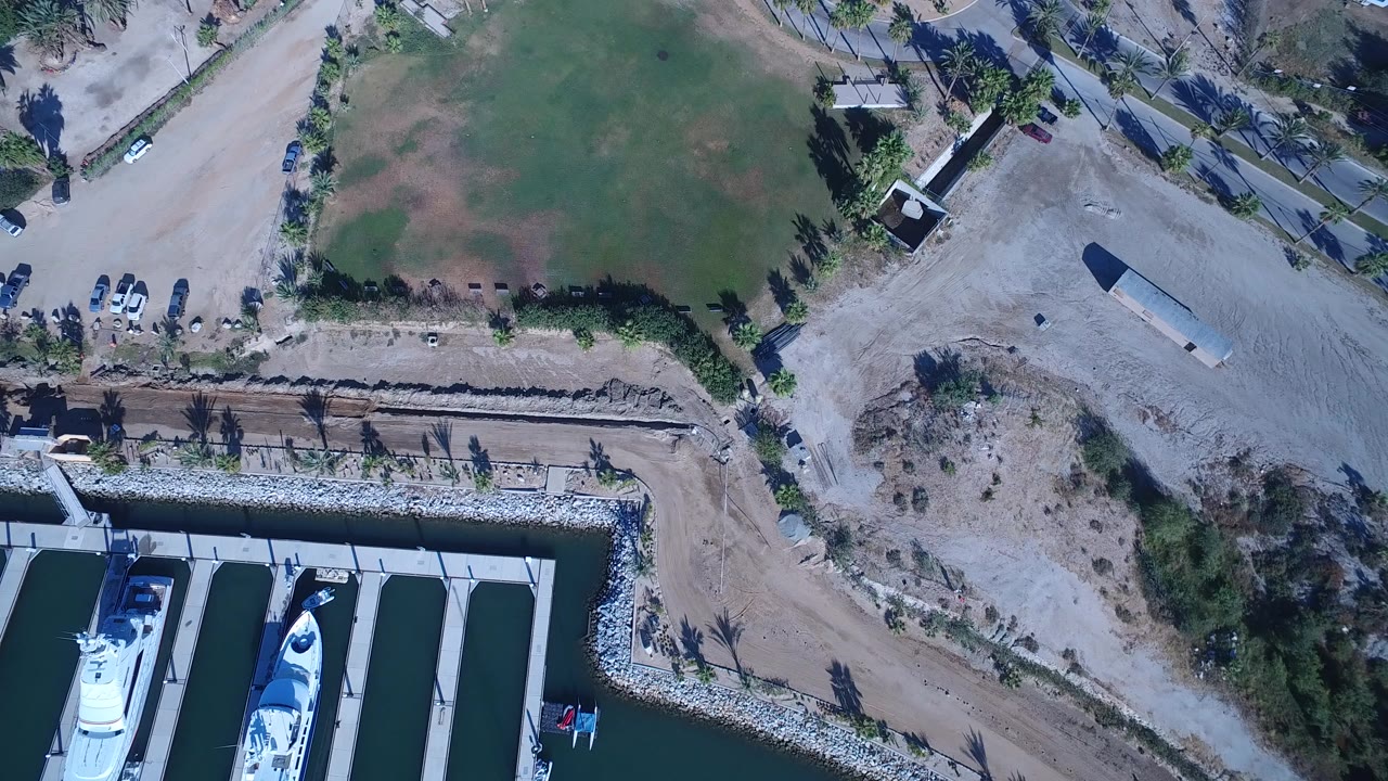 Overhead marina edge with yachts, lawn, road, and dusty ground.