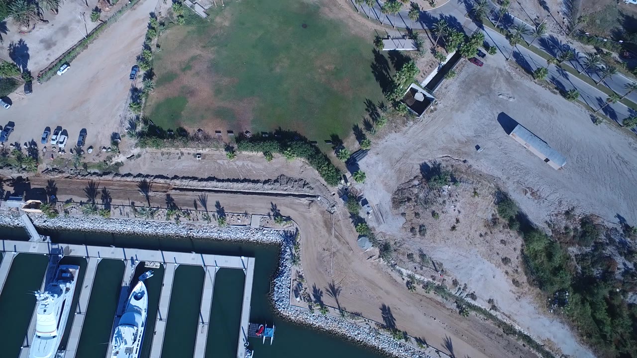 Aerial frame over Playita with two prominent white yachts in marina slips, a rock-lined bank, a patchy green field, pale dirt roads, parked vehicles, palms, and very small possible people near the road edge.