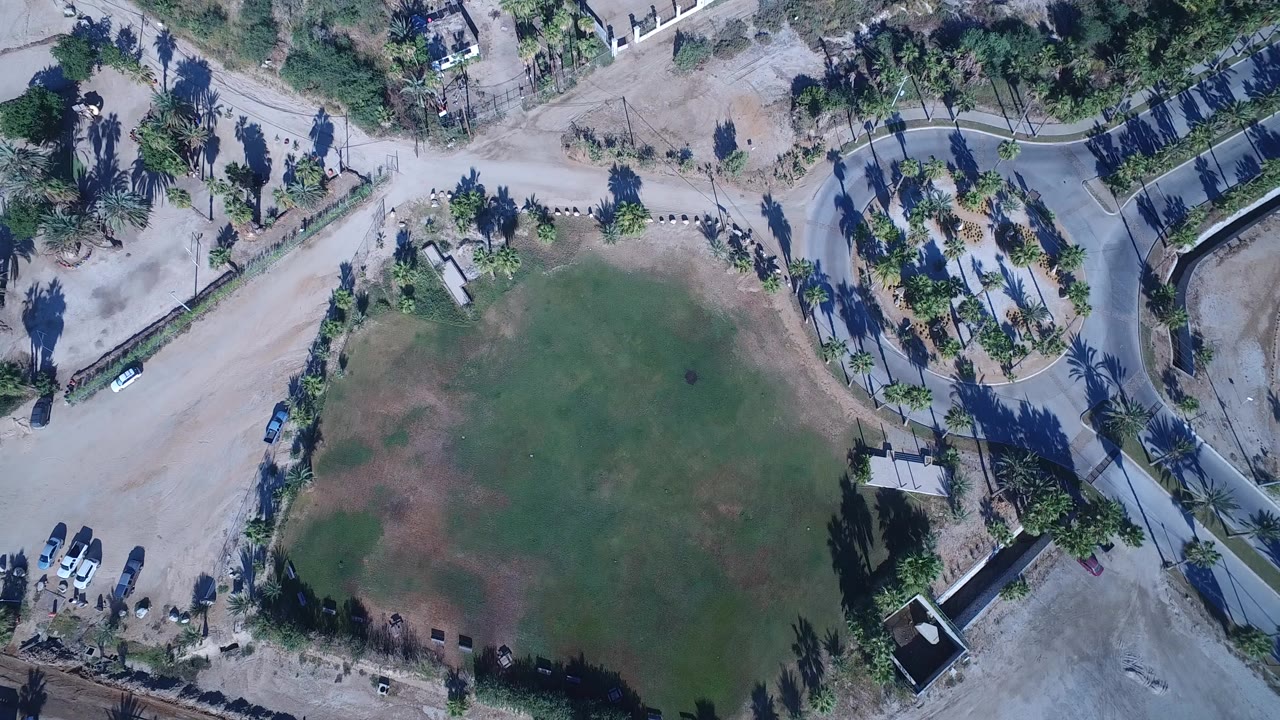 High aerial view over Playita, Baja California Sur, with a patchy green field, curving road, dirt lots, palm shadows, small houses, parked cars, and no visible people.