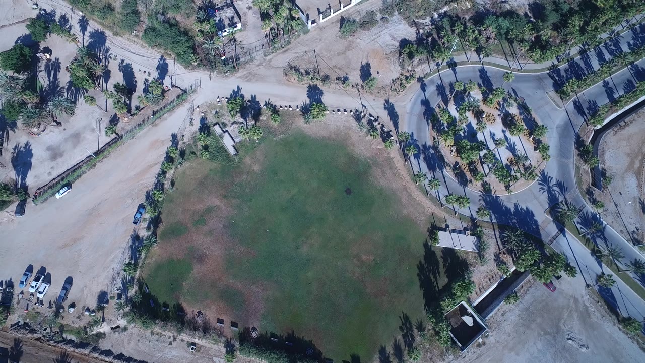 Overhead Playita frame showing the green field lower in view, pale dirt road sections, palms, small buildings, parked vehicles, garden walls, and no visible people.