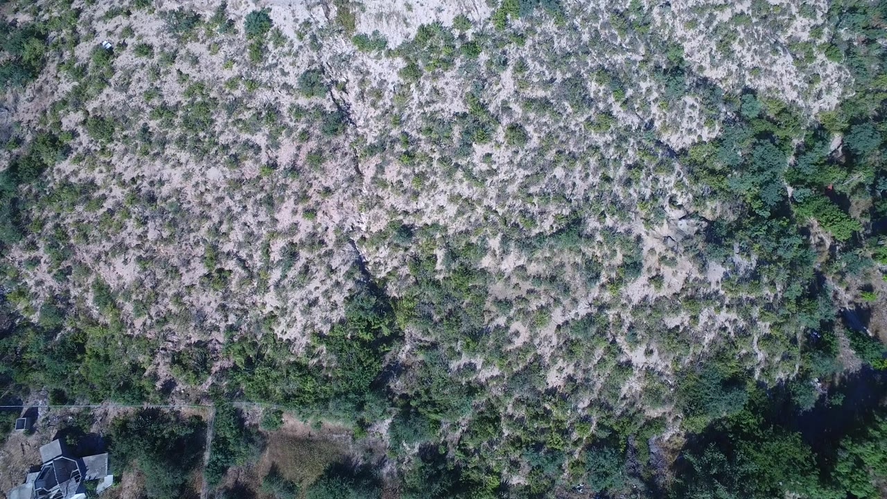 Aerial frame over Playita scrubland with mottled pale ground, dense green vegetation pockets, small edge structures near the lower frame, and no visible people.