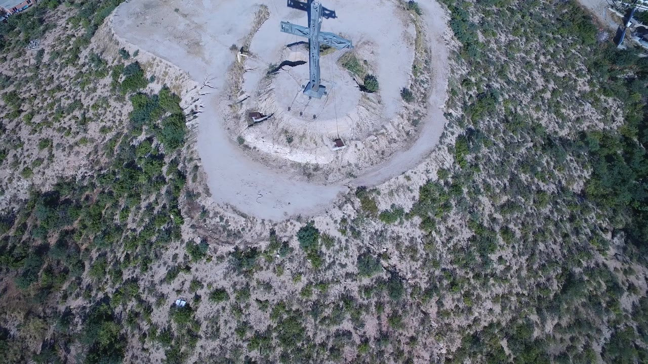 Aerial frame showing the cross statue and its dark shadow stretching across pale sand in Playita, with dense scrub vegetation filling the right side and no visible people.