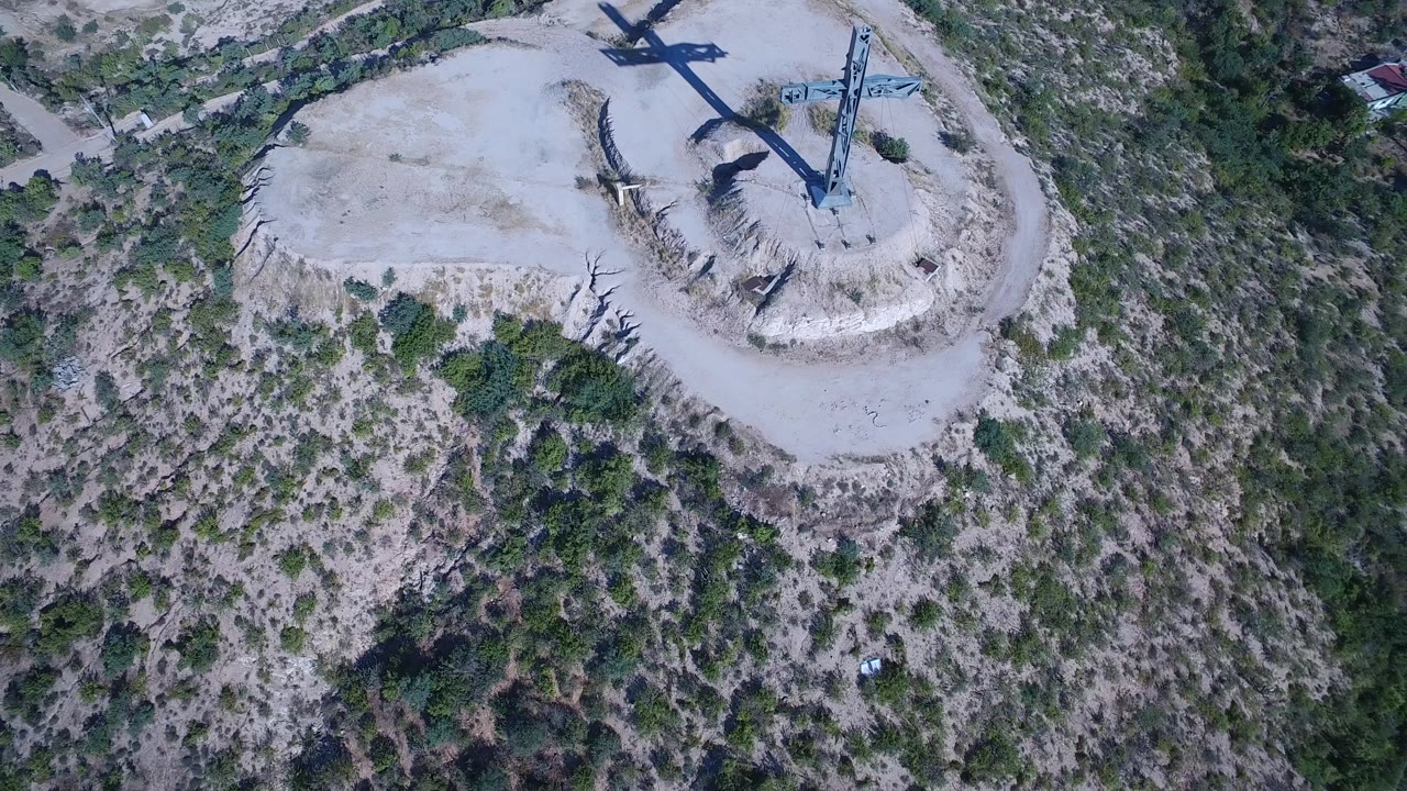 Aerial frame from Playita, Baja California Sur with the steel cross casting a dark form across the sandy hill while dense green brush fills the lower half, with no discernible faces.