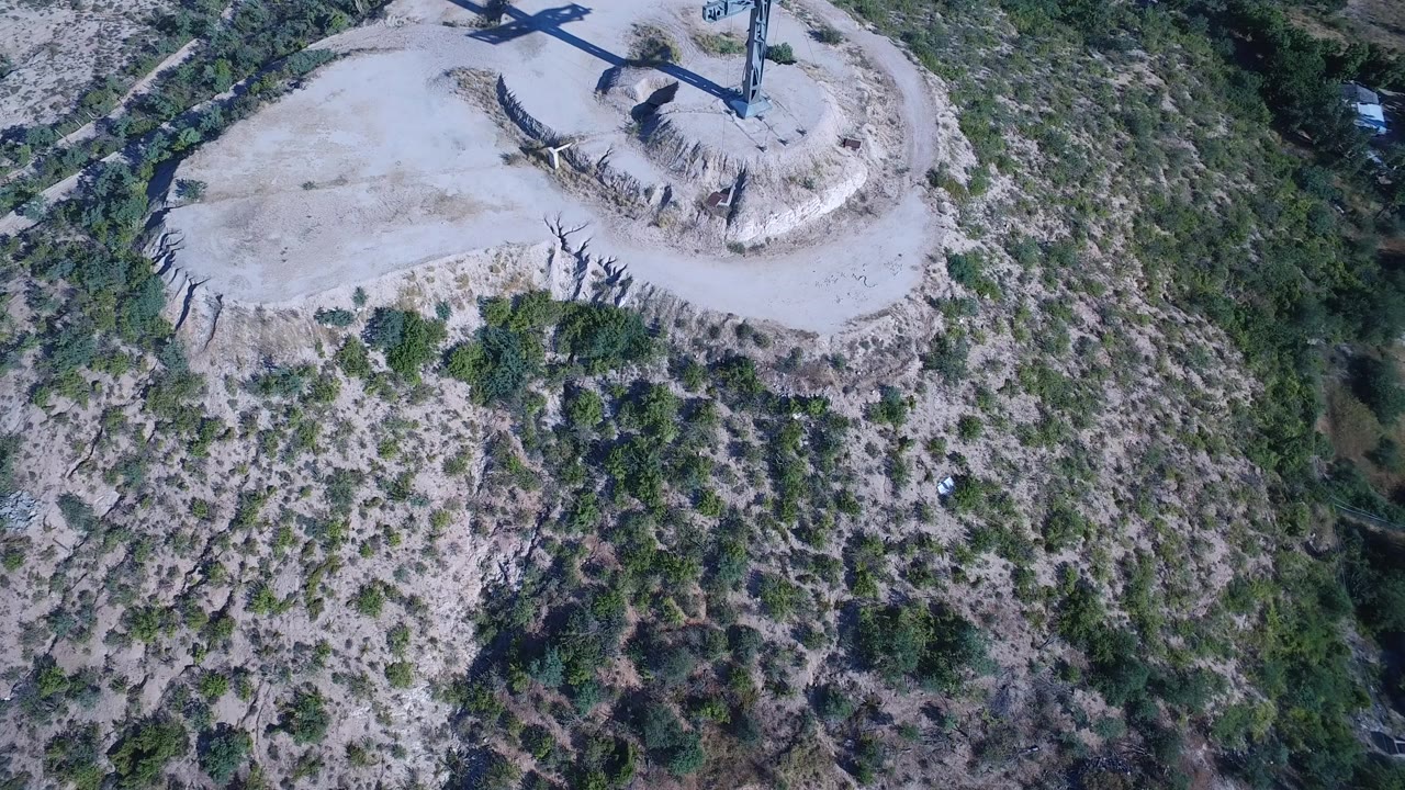 Opening aerial frame over Playita, Baja California Sur, showing a large cross near the top edge of a pale rocky hill with scrub-covered slopes below and no visible people.