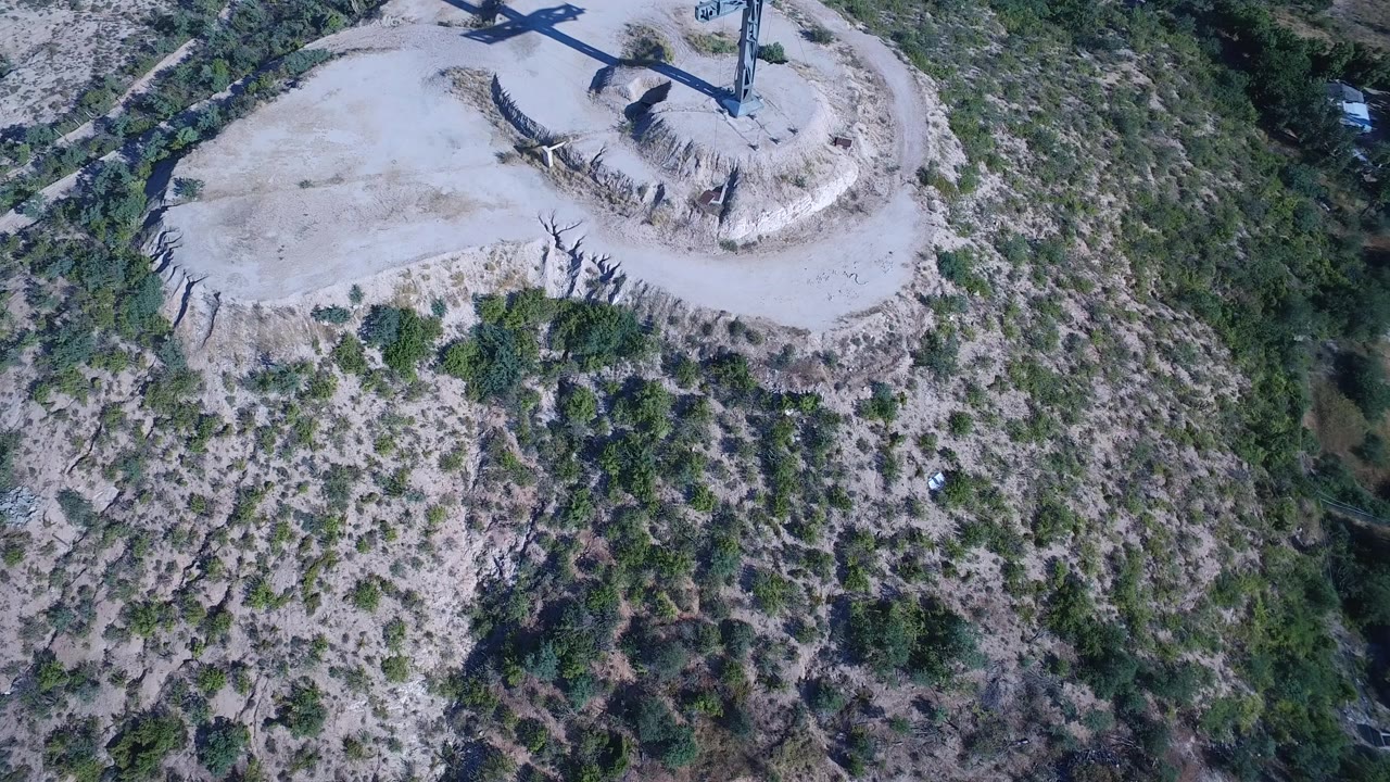 Aerial view in Playita, Baja California Sur, with the cross and its dark shadow over the cleared hilltop while rough green scrub fills the lower frame and no visible people appear.