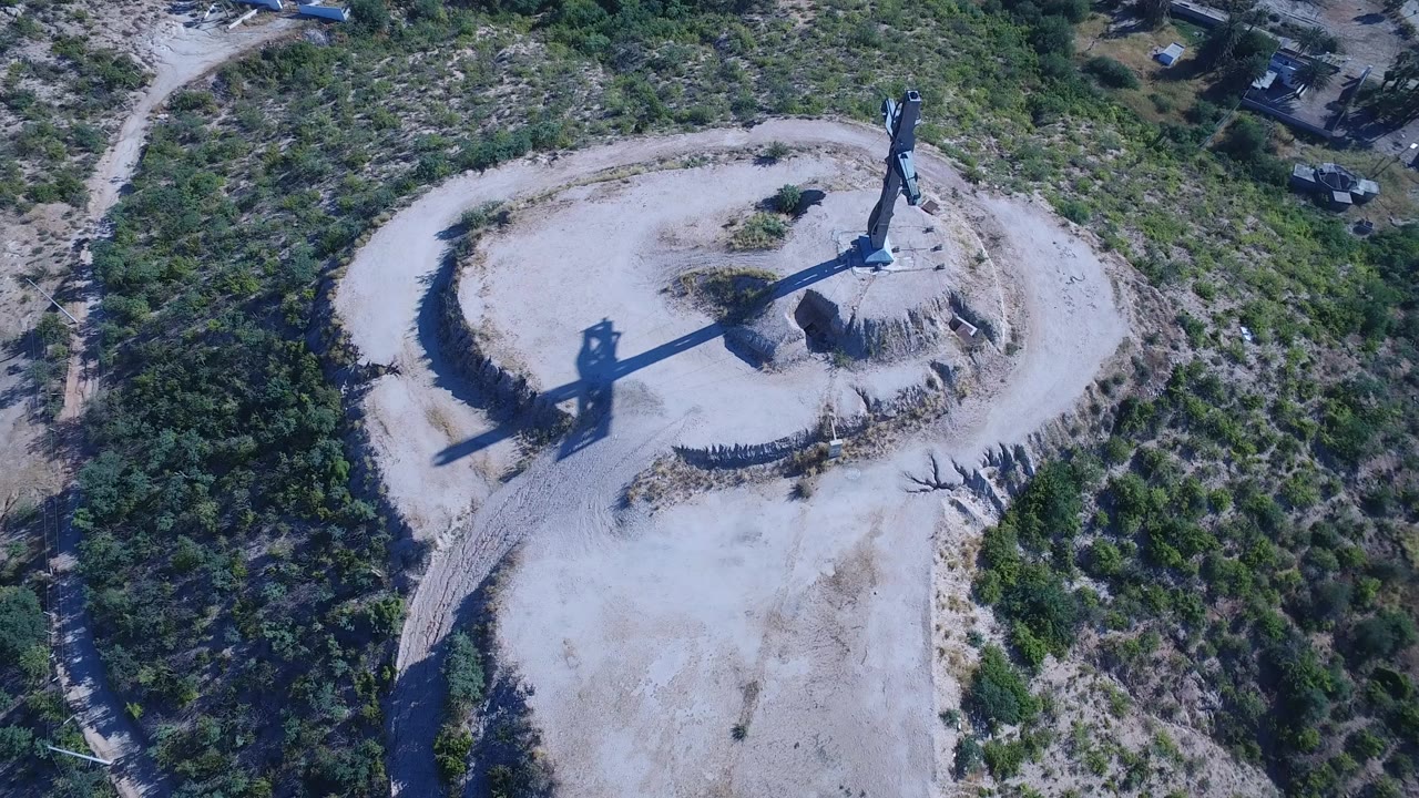 Aerial view of a circular dirt clearing with a tall sculptural structure and scrubland around it.