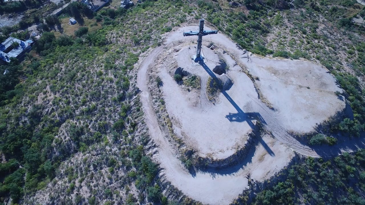 Overhead view of a hilltop cross on a pale dirt mound surrounded by scrub vegetation.