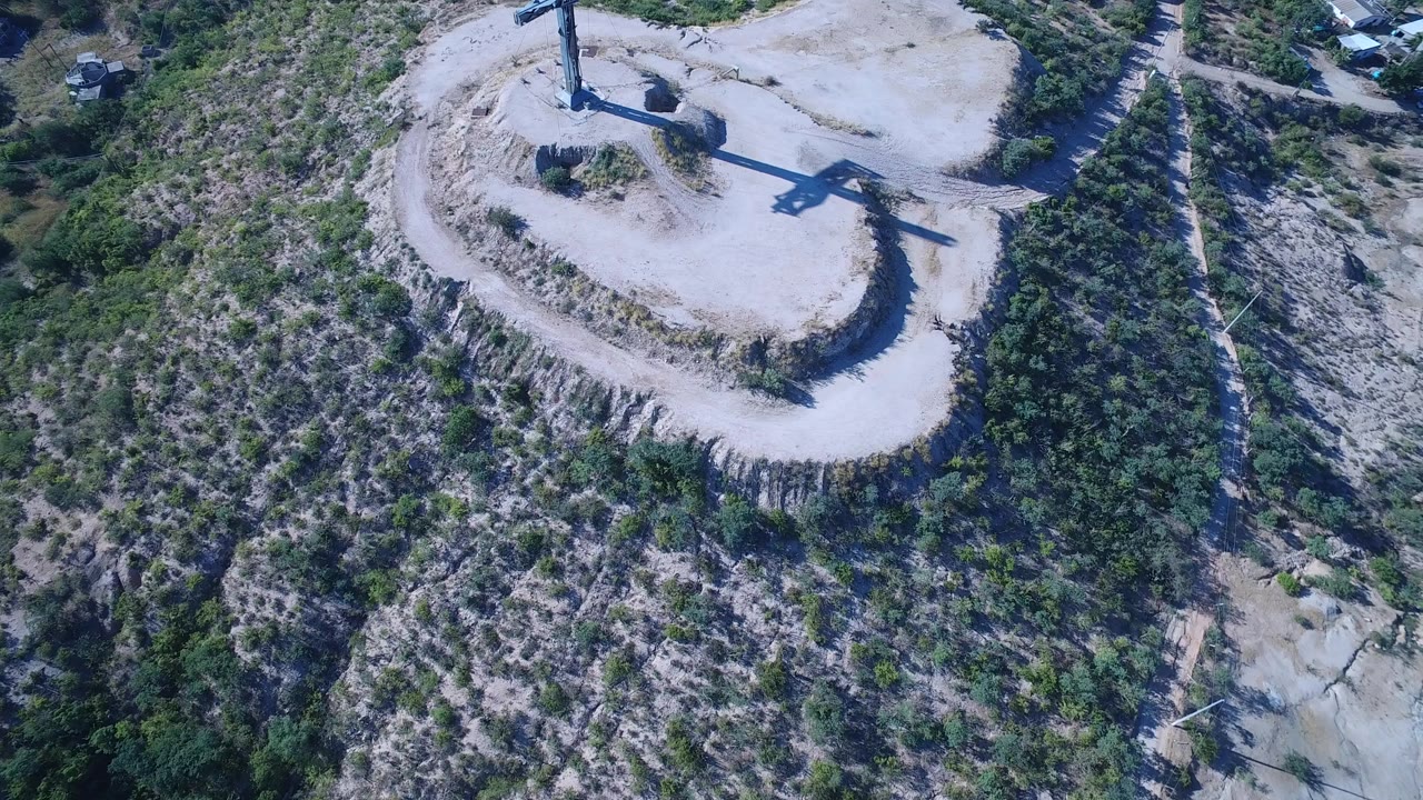 Opening aerial frame in Playita, Baja California Sur shows a tall crucifix on a pale hilltop beside a curving dirt road, with scrub-covered slopes below and no visible people.