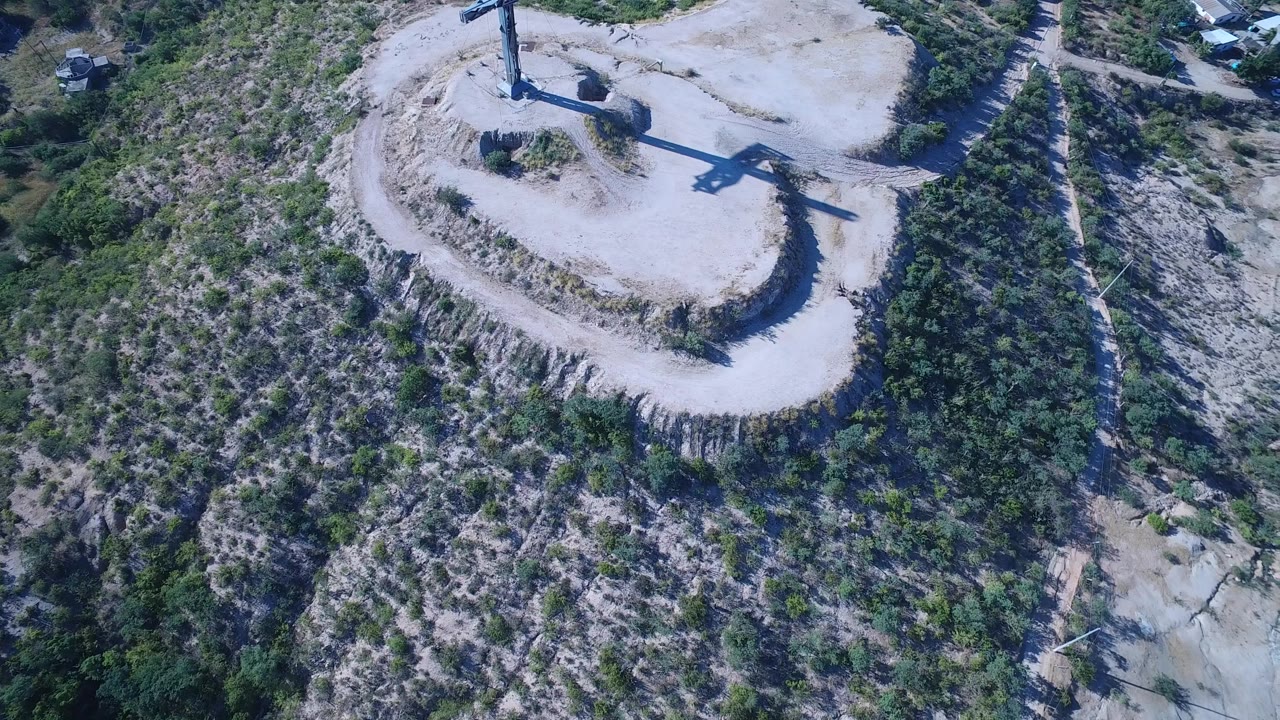 A slightly later aerial view in Playita, Baja California Sur keeps the crucifix near the top of frame, with its shadow crossing the pale ground and no visible people.
