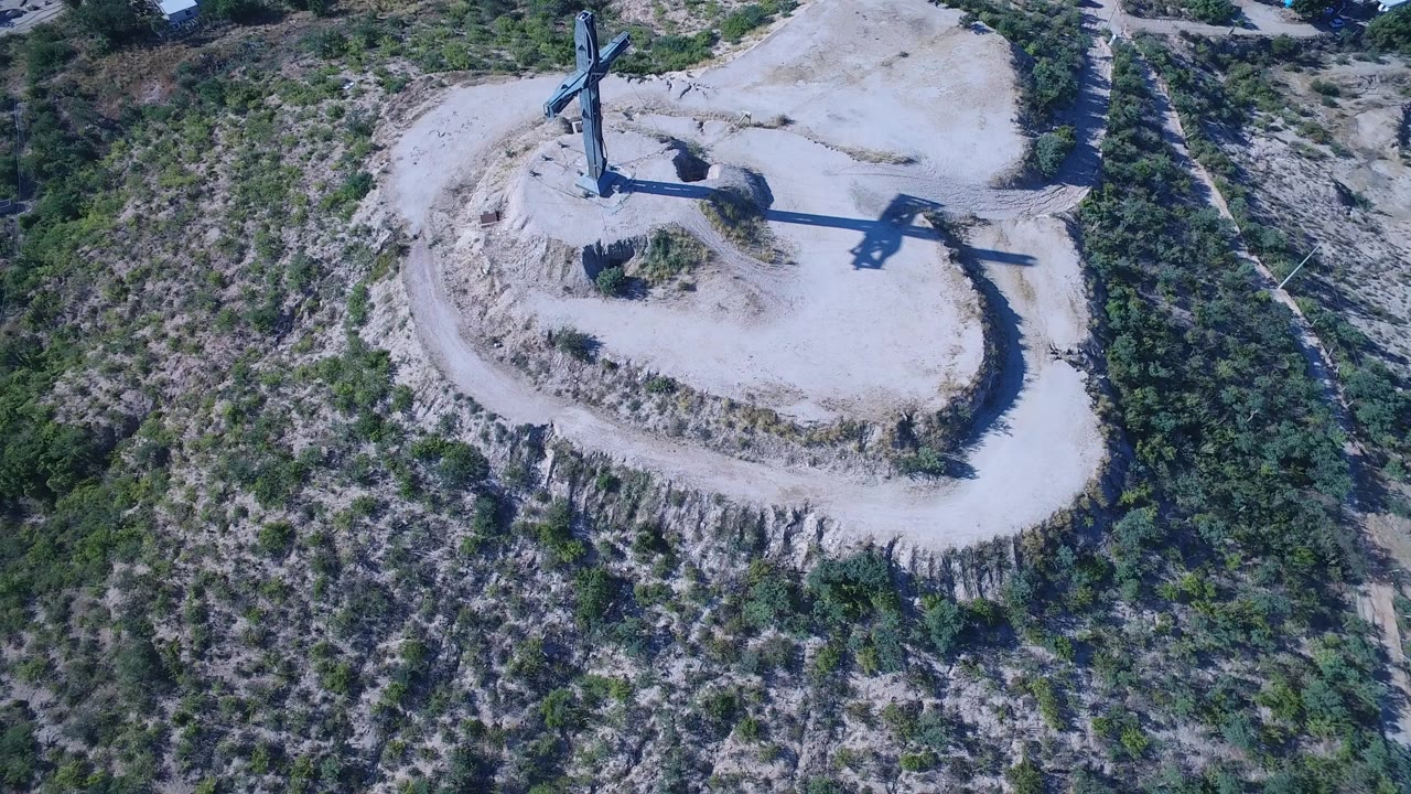 Aerial view of a cross on a pale dirt hilltop surrounded by scrubland.