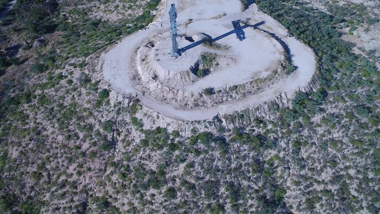 High aerial frame over Playita, Baja California Sur showing a blue cross near the top of a pale dirt mound, dry scrubland around it, and no visible people.