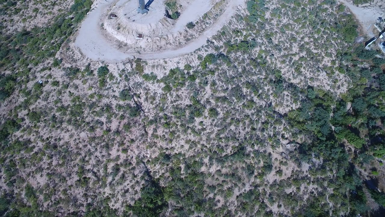 Overhead drone frame in Playita, Baja California Sur, Mexico showing a pale dirt loop and cross near the upper edge above rocky scrubland, with no visible people.