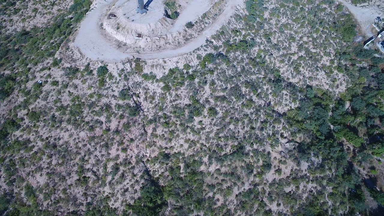 Aerial frame over Playita, Baja California Sur, Mexico with the hilltop cross close to the upper border and rocky green scrub covering the slope below, with no visible people.