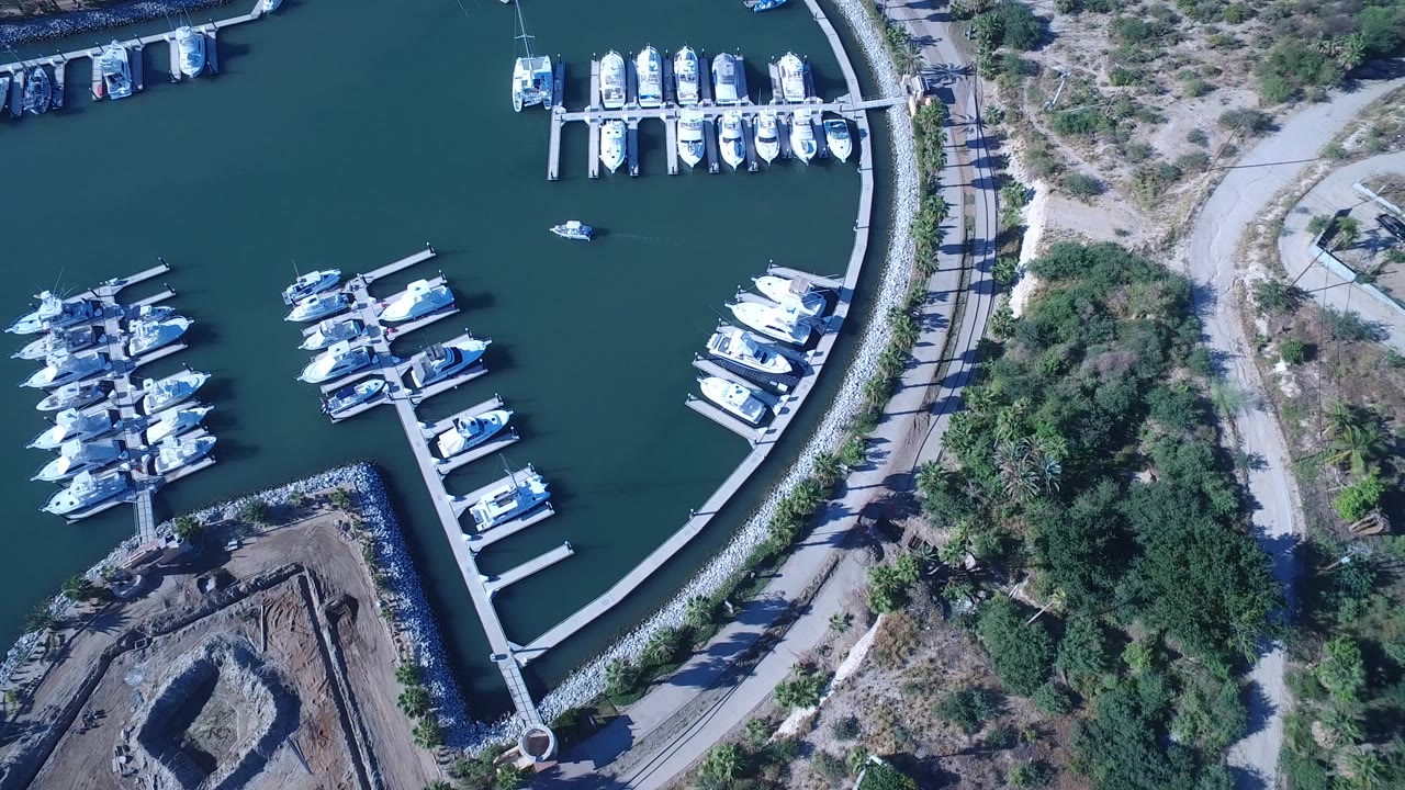 Overhead view of white boats moored in rows at a Playita, Baja California Sur marina, with teal water, docks, a road, trees, and no visible people.