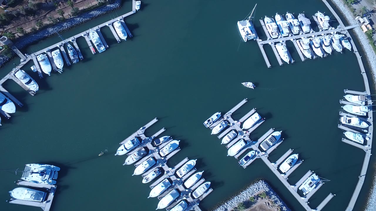 Top-down aerial frame over Playita, Baja California Sur showing white boats moored along docks in blue-green water, with rock shoreline edges and no visible people.